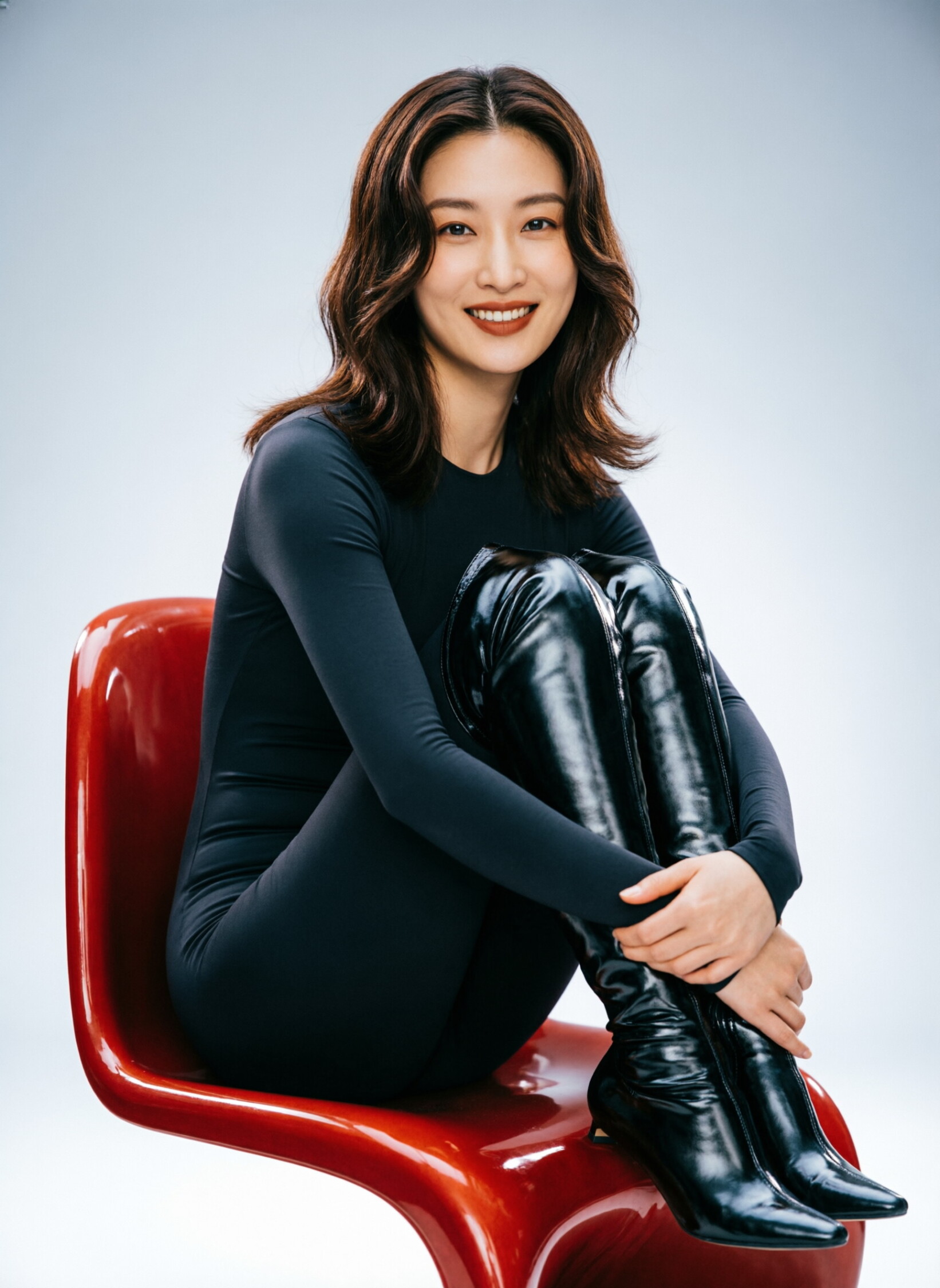 Seated facing forwards in three-quarter profile on a red shiny modern designer chair with the knees drawn toward the chest